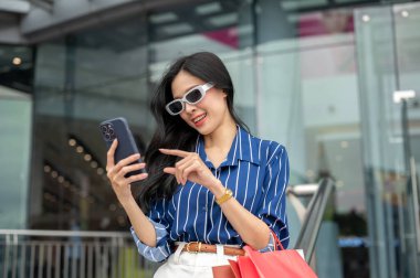 Güneş gözlüklü Asyalı kadın alışveriş merkezinin önünde elinde çantayla telefona bakıyor. Maaş günü, tanıtım reklamı, modern şehir hayatı..
