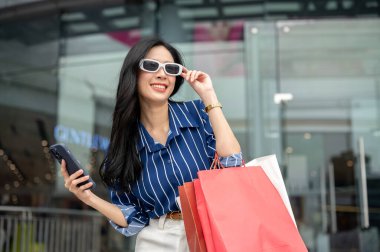 Güneş gözlüğü takan Asyalı kadın alışveriş merkezinin önünde elinde çantalarla duruyor. Maaş günü, tanıtım reklamı, modern şehir hayatı..