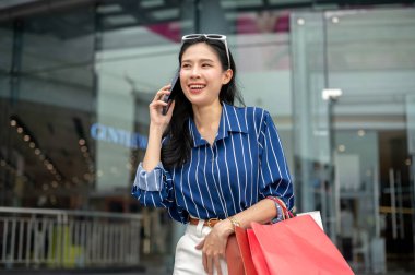 Alışveriş merkezinin önünde alışveriş torbaları taşıyan Asyalı bir kadın elinde telefonla konuşuyor. Maaş günü, tanıtım reklamı, modern şehir hayatı..