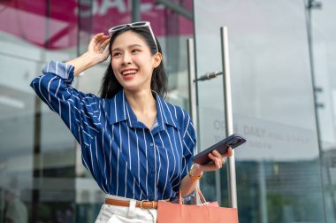 Bir alışveriş merkezinin cam kapısından çıkarken elinde alışveriş çantası olan Asyalı bir kadın. Maaş günü, tanıtım reklamı, modern şehir hayatı..