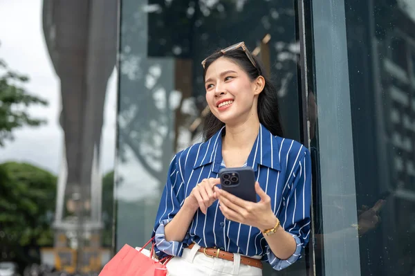 Alışveriş çantası taşıyan ve elinde telefonla alışveriş merkezinin önünde duran Asyalı güzel bir kadın. Maaş günü, tanıtım reklamı, modern şehir hayatı..