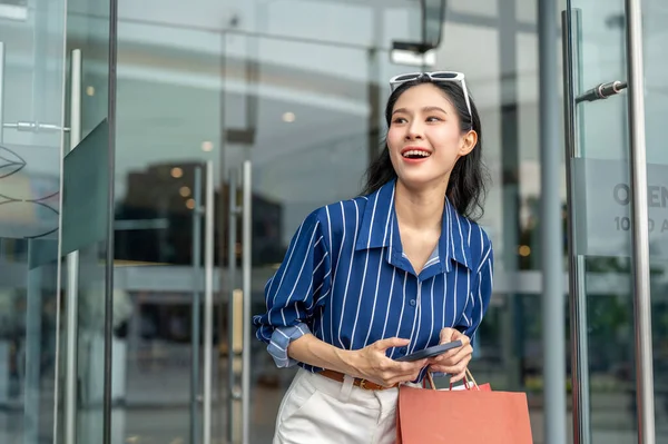 Asyalı kadın elinde telefonla alışveriş merkezinin cam kapısından çıkarken ya da ayaktayken alışveriş çantası taşıyor. Maaş günü, tanıtım reklamı, modern şehir hayatı..