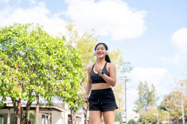 Spor kıyafetli Asyalı bir kadın, kulaklıkla müzik dinlerken güneşli parkta koşu egzersizi yapıyor. Spor yapıyor, sağlıklı yaşıyor, dışarıda takılıyor..