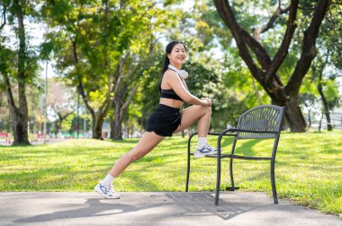 Spor kıyafetli Asyalı bir kadın parkta kaldırımda dururken egzersiz yapmadan önce yedek kulübesinde bacak esnetiyor. Spor yapıyor, sağlıklı yaşıyor, dışarıda takılıyor..