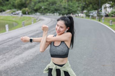 Spor kıyafetli Asyalı bir kadın parkta sokak yolunda dikilirken egzersiz yapmadan önce kollarını esnetiyor. Spor yapıyor, sağlıklı yaşıyor, dışarıda takılıyor..