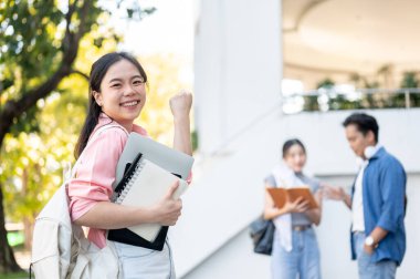 Asyalı bir kız öğrenci dizüstü bilgisayar ve kitap tutuyor. Üniversitede arkadaşlarıyla konuşurken yumruk atıyor. Üniversite, Kampüs Hayatı, Modern Teknoloji.