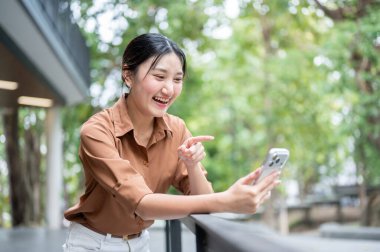 Parkta korkuluklara yaslanırken akıllı telefonu tutan ve işaret eden Asyalı kız öğrenci. Boş zaman, Kolej Kampüsü, Şehir Hayatı.
