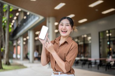 Asyalı bir kız öğrenci açık hava binasında dikilirken beyaz ekrandaki akıllı telefonu işaret ediyor. Boş zaman, Kolej Kampüsü, Şehir Hayatı.