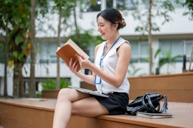 Asyalı ofis çalışanı kadın parkta tahta merdivenlerde bacak bacak üstüne atarak kitap okuyor. Çalışan Kartı, Modern Şirket, Serinletici Açık Hava.