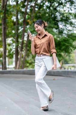 Parkta merdivenlerde yürürken elinde dizüstü bilgisayarla telefona bakan mutlu Asyalı kız öğrenci. Boş zaman, Kolej Kampüsü, Şehir Hayatı.