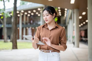 Açık hava binasında ayakta dururken ya da yürürken tablete bakan Asyalı bir kız öğrenci. Boş zaman, Kolej Kampüsü, Şehir Hayatı.