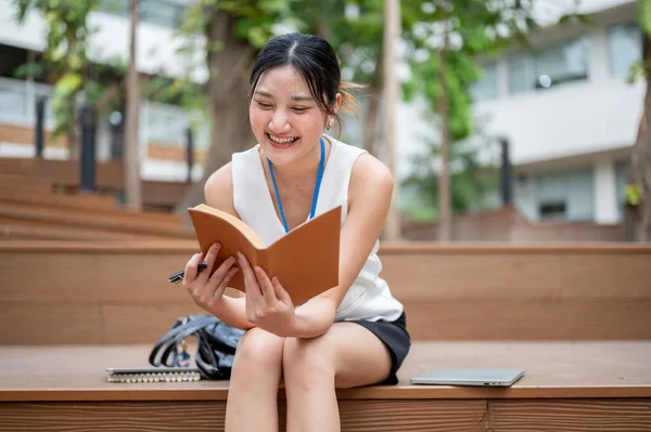 Asyalı ofis çalışanı kadın elinde kalemle kitap okurken parktaki tahta merdivenlerde çantasıyla oturuyor. Çalışan Kartı, Modern Şirket, Şehir Hayatı.