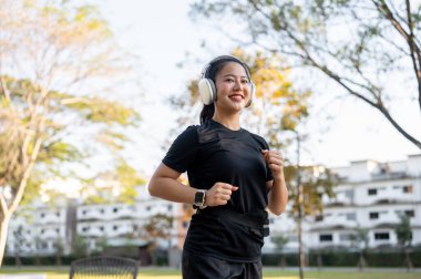 Spor kıyafetli Asyalı bir kadın parkta koşarken kulaklıkla müzik dinliyor. Sağlıklı Yaşam, Spor, Kentsel Modern Yaşam