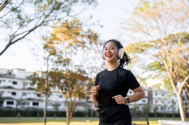 Spor kıyafetli Asyalı bir kadın parkta koşarken kulaklıkla müzik dinliyor. Sağlıklı Yaşam, Spor, Kentsel Modern Yaşam