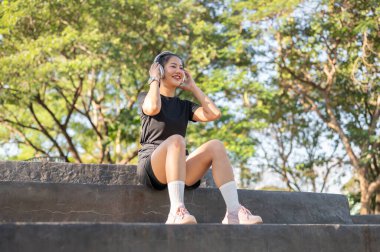 Spor kıyafetli, elinde kulaklıkla müzik dinleyen Asyalı bir kadın parkta egzersiz yaptıktan sonra merdivenlerde oturuyor. Sağlıklı Yaşam, Spor, Kentsel Modern Yaşam