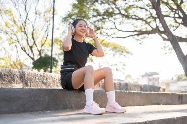 Siyah spor kıyafetli Asyalı kadın parkta egzersiz yaptıktan sonra beton merdivenlerde oturan kulaklık takıyor. Sağlıklı Yaşam, Spor, Kentsel Modern Yaşam