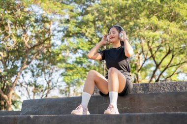 Spor kıyafetli Asyalı kadın parkta egzersiz yaptıktan sonra merdivenlerde kulaklık müziği dinlerken gözlerini kapadı. Sağlıklı Yaşam, Spor, Kentsel Modern Yaşam