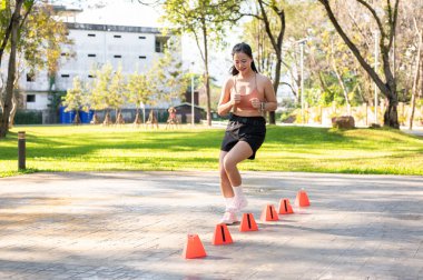 Spor kıyafetleri içinde, güneş ışığında park kaldırımında koni talimi yapan Asyalı güzel bir kadın. Sağlıklı Yaşam, Spor, Kentsel Modern Yaşam