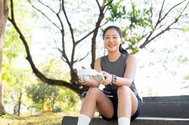 Spor kıyafetli gülümseyen Asyalı kadın elinde kulaklıkla yeşil bahçe parkında beton merdivenlerde oturuyor. Sağlıklı Yaşam, Spor, Kentsel Modern Yaşam