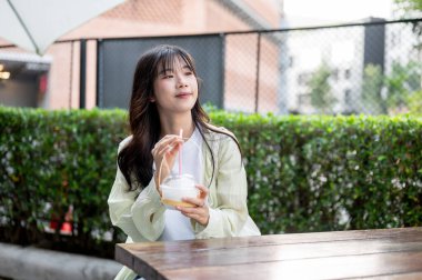 Mutlu Asyalı kadın turist elinde samanla alışveriş merkezinde oturmuş hindistan cevizi suyu içiyor. Tek başına yolculuk, şehir turu, açık havada dinlenme..