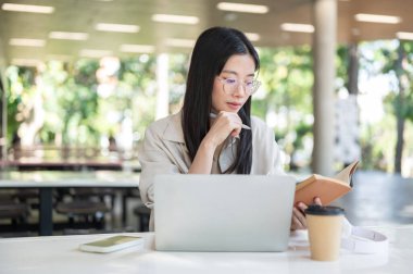 Beyaz masada elinde kalem okuma kitabı tutan Asyalı gözlüklü bir üniversite öğrencisi. Ödev işi, Üniversite Kampüsü, Şehir Hayatı.