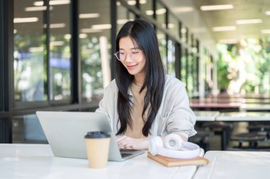 Gözlüklü Asyalı üniversite öğrencisi. Binada otururken dizüstü bilgisayara bakıyor ya da daktilo kullanıyor. Boş zaman, Üniversite Kampüsü, Şehir Hayatı.