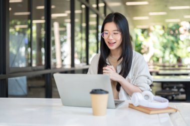 Gözlüklü Asyalı bir üniversite öğrencisi laptopa binadaki masada oturuyormuş gibi bakıyor. Boş zaman, Üniversite Kampüsü, Şehir Hayatı.