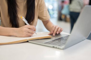 Binanın masasında dizüstü bilgisayarla yazı yazarken elinde kalem tutan kadın üniversite öğrencisinin yakın çekimleri. Ödev işi, Üniversite Kampüsü, Şehir Hayatı.