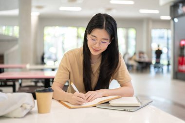 Gözlüklü kolej öğrencisi elinde kalemle dizüstü bilgisayarının yanında binadaki masada yazıyor. Ödev işi, Üniversite Kampüsü, Şehir Hayatı.