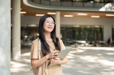 Dizüstü bilgisayar taşıyan Asyalı gözlüklü bir üniversite öğrencisi. Binada ayakta ya da yürüyor. Boş zaman, Üniversite Kampüsü, Şehir Hayatı.