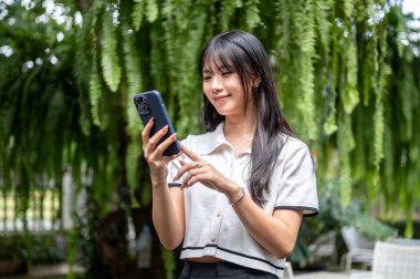 Cafe 'nin bahçesindeki ağacın altında dururken akıllı telefonu tutan ve ona bakan güzel Asyalı kadın. Boş zaman, estetik mekan, Serinletici Açık Hava.