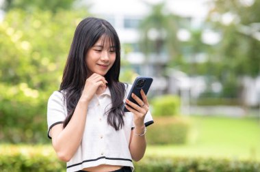 Asyalı güzel bir kadın, yeşil bahçe kafesinde ya da parkta dururken telefona bakıyor. Boş zaman, estetik mekan, Serinletici Açık Hava.