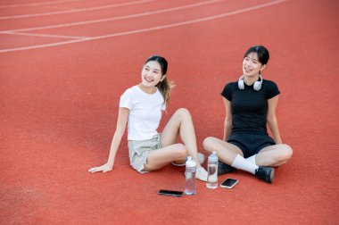 Asyalı bir kadın ve arkadaşı spor stadyumunda pistin kenarında otururken etrafa bakınıyorlar. Spor, sağlıklı yaşam, açık hava aktivitesi.