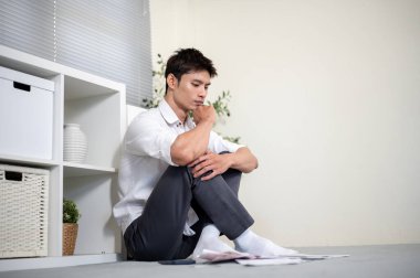 Asyalı ofis çalışanı çenesini tutarak oturma odasında otururken faturalara bakar gibi düşünüyor. Finansal Planlama, Para Yönetimi, Şehir Hayatı.