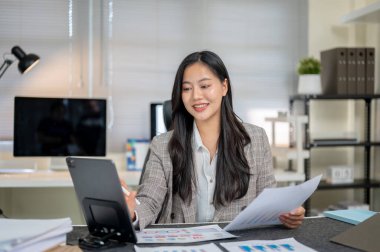Mutlu iş kadını ya da ofis çalışanı elinde belge tutarak tablete şirket masasında oturuyormuş gibi bakıyor. Masa başı işi, çalışma saatleri, şirket çalışanı..