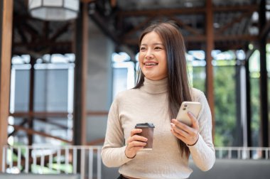 Süveter giymiş gülümseyen Asyalı kadın kafeteryada yürürken elinde kahve ile telefonundan başka tarafa bakıyor. Boş Zaman, Modern Teknoloji, Dışarıdaki Dinlenme.