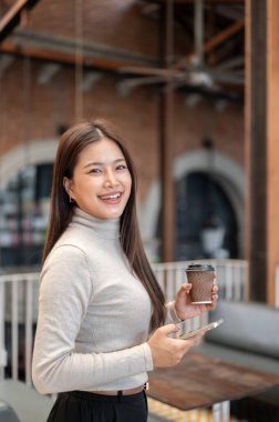 Gülümseyen Asyalı kadın kahve fincanı ve akıllı telefon tutarken yan yana dururken ya da kafede yürürken. Boş Zaman, Modern Teknoloji, Dışarıdaki Dinlenme.