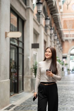 Elinde kahve ve telefon olan Asyalı güzel bir kadın alışveriş merkezinde yürüyormuş gibi yukarı bakıyor. Boş zaman, Solo gezi, Seyahat Turisti.