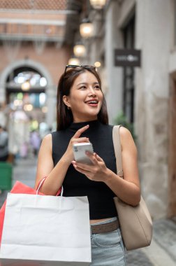 Güzel Asyalı bir kadın elinde telefon ve alışveriş torbalarıyla alışveriş merkezinde dikilirken yukarı bakıyor. Boş zaman, Solo Seyahat, Turist Satın Alma.