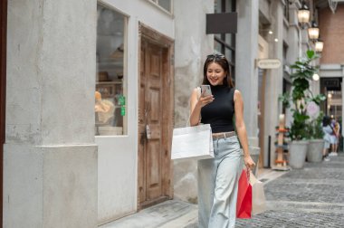 Alışveriş torbaları taşıyan ve alışveriş merkezinde ya da markette yürürken telefona bakan mutlu Asyalı kadın. Boş zaman, Solo Seyahat, Turist Satın Alma.