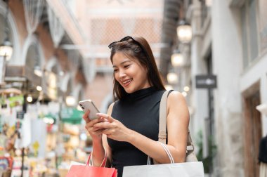 Mutlu Asyalı kadın telefonuna bakıyor ve alışveriş merkezinde yürürken ya da dikilirken alışveriş torbalarını tutuyor. Boş zaman, Solo Seyahat, Turist Satın Alma.