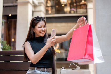 Alışveriş torbaları tutan mutlu Asyalı kadın telefonla fotoğraf çekiyor. Alışveriş merkezindeki ahşap bankta oturuyor. Eğlence Zamanı, Seyahat Gezisi, Müşteri Alımı.
