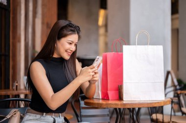 Güzel Asyalı bir kadın kafede alışveriş torbalarıyla otururken telefonda konuşuyor ya da bakıyor. Eğlence Zamanı, Seyahat Gezisi, Müşteri Alımı.