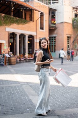 Alışveriş merkezinde alışveriş torbalarıyla yürürken telefonu tutan güzel Asyalı kadın arkasını dönüyor. Boş Zaman, Seyahat Gezisi, Turist Müşteri.