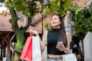 Alışveriş merkezinde ayakta ya da yürürken akıllı telefon taşıyan Asyalı güzel bir kadın. Boş zaman, Solo Seyahat, Turist Satın Alma.