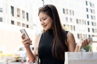 Güzel Asyalı kadın ayakta dururken ya da sokakta yürürken telefona bakıyor ve alışveriş torbaları taşıyor. Boş zaman, Solo Seyahat, Turist Satın Alma.