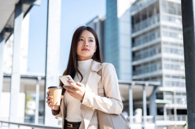 Asyalı kadın ofis çalışanı elinde telefon ve kahveyle yaya köprüsünde yürüyormuş gibi duruyor. Yolda, Günlük rutin, Şehir Modern Hayatı