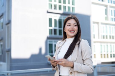 Bej ceketli güzel bir Asyalı kadın ayakta dikilirken ya da yürürken telefonunu kaçırıyor. Yolda, Günlük rutin, Şehir Modern Hayatı