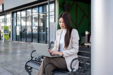 Kaldırımdaki metal bankta bacak bacak üstüne atarken bej ceketli güzel Asyalı kadın dizüstü bilgisayar kullanıyor. Şehir Hayatı, Modern Teknoloji, Ürpertici Açık Hava.