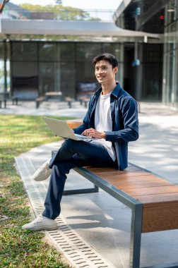 Bahçe parkında tahta bankta bağdaş kurmuş otururken dizüstü bilgisayardan yüzünü çeviren gülümseyen Asyalı adam. Boş Zaman, Modern Teknoloji, Şehir Hayatı.